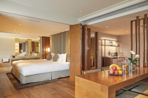 A modern hotel room with a large bed, a wooden desk, and a breakfast bar with fruit.