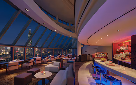 A modern hotel lobby with a high ceiling, large windows offering a city view, and a bar area with seating.