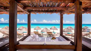 A beach cabana with a wooden roof, white cushioned seating, and a view of the ocean.