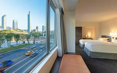 A hotel room with a city view, two beds, a bench, and a window overlooking a busy street.