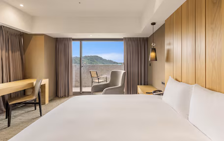 A hotel room with a bed, a desk, a chair, a nightstand, and a view of the outdoors.