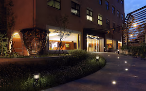 A modern building with a well-lit entrance, a covered outdoor seating area, and a pathway leading to the entrance.