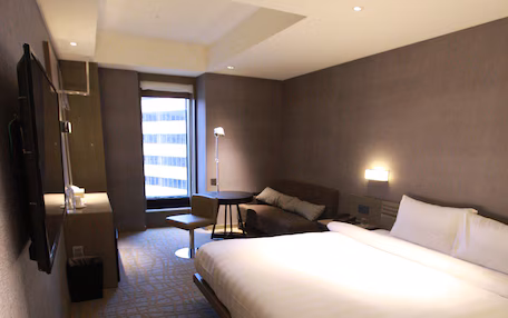 A hotel room with a large bed, a desk, a chair, a TV, and a window with a view of buildings.