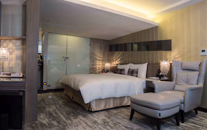 A modern hotel room with a bed, a sitting area with a chair, a desk with a lamp, and a large mirror.