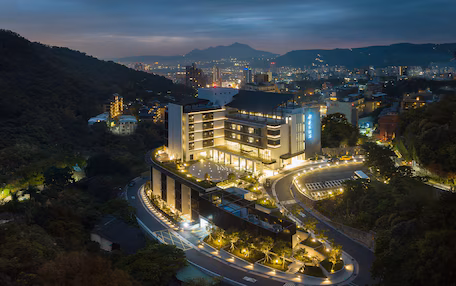 A modern hotel with a curved driveway, surrounded by greenery and a mountainous backdrop.