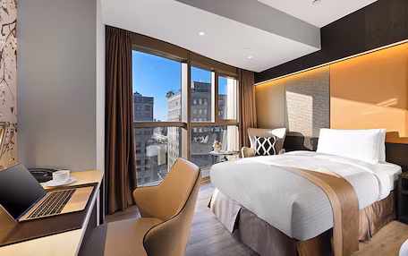 A modern hotel room with a large bed, a desk with a laptop, a chair, and a view of the city through large windows.