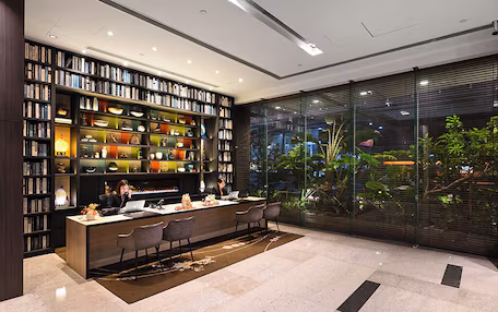 A modern library with bookshelves, a reception desk, and a view of greenery through large windows.