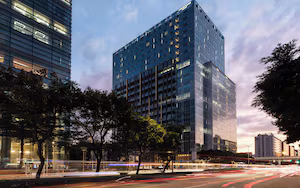 A modern high-rise building with a glass facade, surrounded by trees and a busy street with light trails.