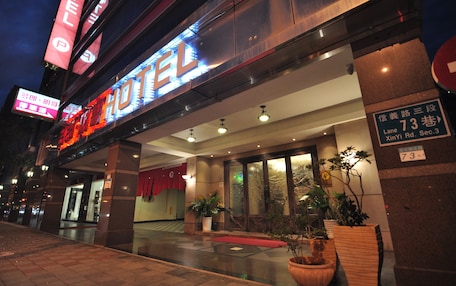 A hotel entrance with a neon sign, potted plants, and a red carpet.