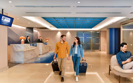 A hotel lobby with a reception desk, a digital sign, a blue patterned floor, and a glass elevator.