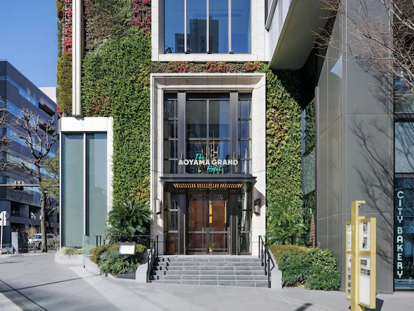 A hotel entrance with a modern facade, a visible staircase, and a sign for 'The Aoyama Grand Hotel.'