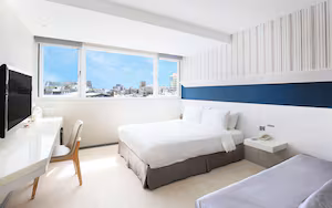 A modern hotel room with a large bed, a desk, and a chair. There is a window with a city view.
