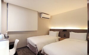 A hotel room with two beds, a desk, a chair, an air conditioning unit, and a wall-mounted lamp.