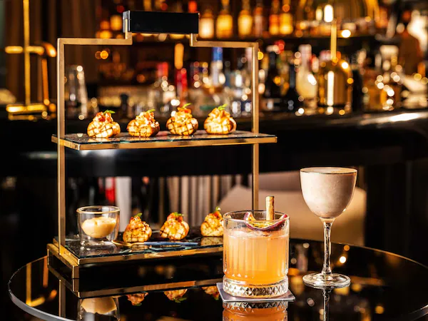 A bar setting with a three-tiered dessert display, a cocktail glass, and a bottle of liquor on a reflective table.