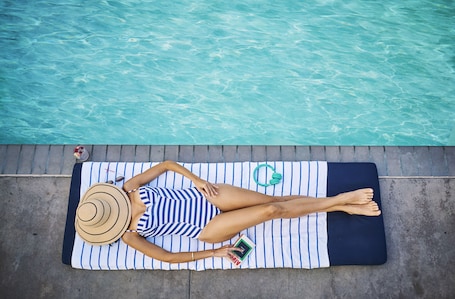 A person lying on a striped towel by a pool, with a book and a green water bottle nearby.