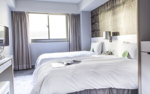 A hotel room with two beds, a large window with curtains, a bookshelf, and a bedside table with a lamp.