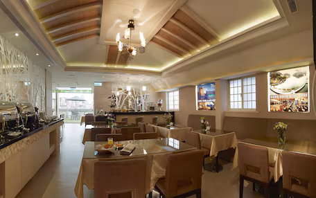 A hotel dining area with a buffet, tables, chairs, and a view of the cityscape through large windows.