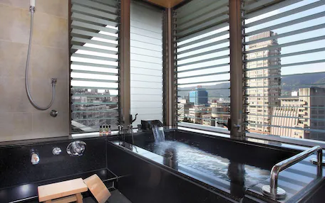 A modern bathroom with a large bathtub, a shower, and a view of the city through large windows.