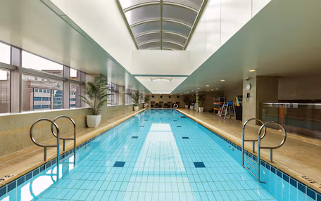 A large indoor swimming pool with clear blue tiles, metal handrails, and potted plants along the sides.