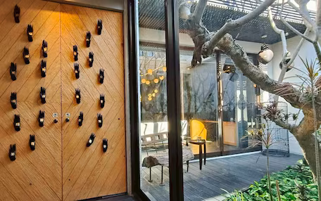 A wooden wall with hand-shaped cutouts, a glass door, and a view of an outdoor seating area with plants and a tree.