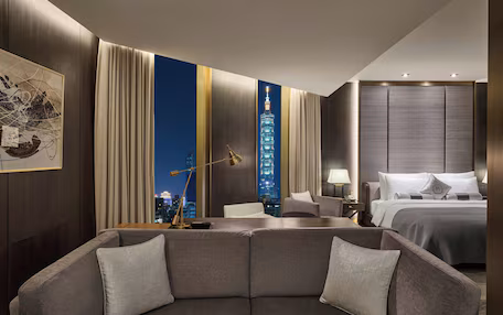 A modern hotel room with a large bed, a seating area with a sofa, a desk, and a view of a cityscape at night.