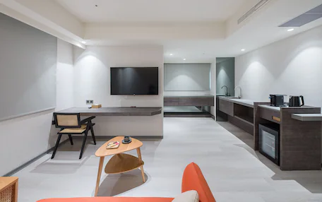 A modern hotel room with a flat-screen TV, a dining area with a table and chairs, a kitchenette, and a sofa.