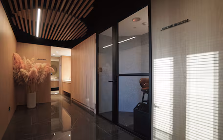 A modern hotel lobby with a wooden reception desk, a glass door leading to a seating area, and a large vase of pampas grass.