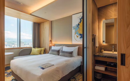 A hotel room with a large bed, a desk, a bathroom with a sink and mirror, and a view of the city.
